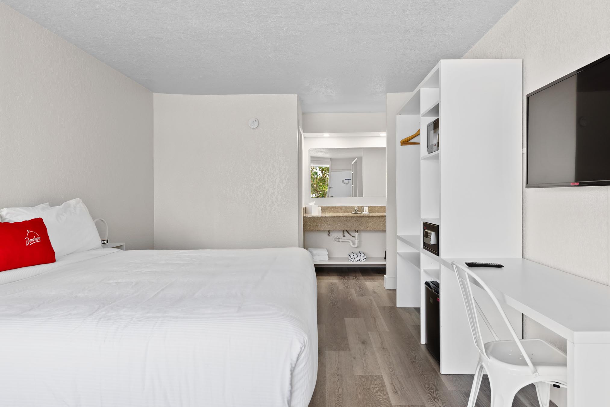 King bed with white sheet, desk, chair, TV mounted on wall and view of bathroom